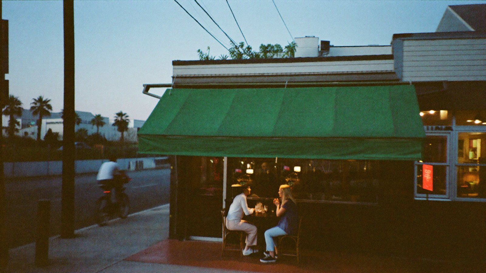 Dusk photograph of the Halloran corner on Grand Avenue — two regulars at a sidewalk table under the green awning, a cyclist passing in motion blur