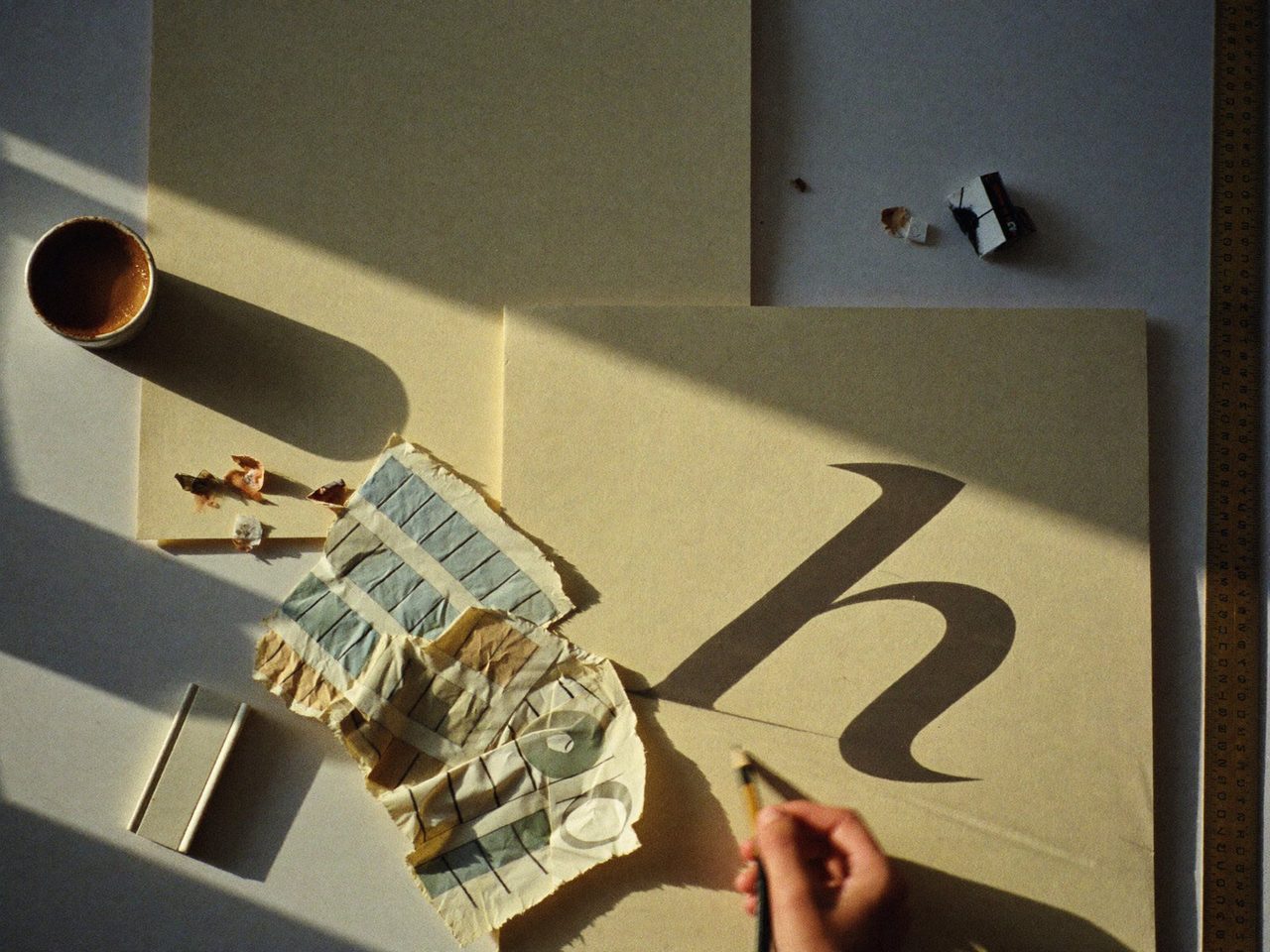 The maker's desk, mid-sketch: a hand drawing a serif letterform on cream paper among paint swatches and afternoon shadows